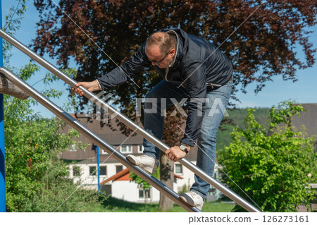 An adult man climbs a metal playground structure on a sunny day. He wears casual clothes and sunglasses, enjoying a moment of physical activity in a public park setting. An adult man climbs a metal playground structure on a sunny day. He wears casual clothes and sunglasses, enjoying a moment of physical activity in a public park setting. 126273161