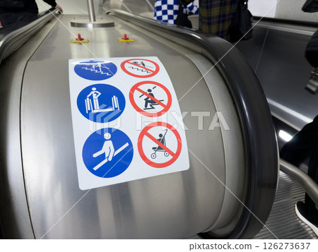 Escalator safety instruction sticker with visible passengers in background, showing real life context of public transport and importance of following safety guidelines Escalator safety instruction sticker with visible passengers in background, showing real life context of public transport and importance of following safety guidelines 126273637