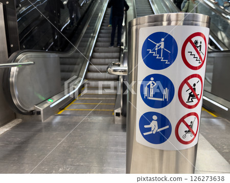 Closeup of escalator safety instruction sign in metro station with visible stairs and people in background, illustrating public space design and visual safety communication 126273638