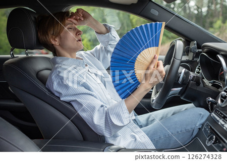 Tired exhausted middle aged woman waving blue fan suffers from stuffiness driving car.  126274328