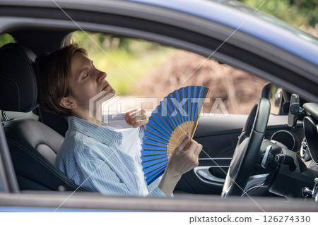 Tired exhausted middle aged woman waving blue fan suffers from stuffiness driving car.  126274330