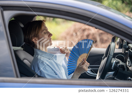 Tired exhausted middle aged woman waving blue fan suffers from stuffiness driving car. Tired exhausted middle aged woman waving blue fan suffers from stuffiness driving car. 126274331