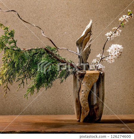 Beautiful spring ikebana. Floral arrangement with spring blooming white flowers, thuja branch and piece of wood in brown ceramic vase, standing on wooden table. Japanese style home decor 126274600