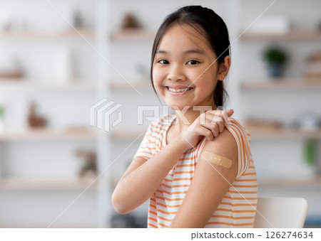 Healthy asian kid girl got vaccinated, sitting on couch at home and showing adhesive band on her shoulder, smiling at camera, panorama with copy space. Kids vaccination, immunization concept Healthy asian kid girl got vaccinated, sitting on couch at home and showing adhesive band on her shoulder, smiling at camera, panorama with copy space. Kids vaccination, immunization concept 126274634