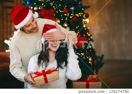 Loving young handsome man giving surprise gift to his wife, closing her eyes, celebrating Christmas at home Loving young handsome man giving surprise gift to his wife, closing her eyes, celebrating Christmas at home 126274780