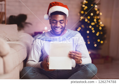 Happy afro man opening xmas box, wearing santa hat and sitting against Christmas Tree at home 126274788
