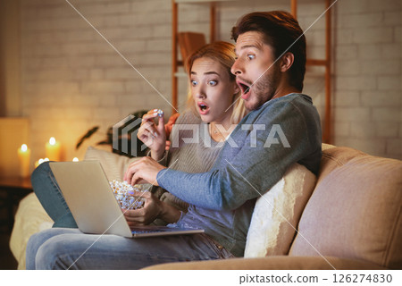 Movie Night. Surprised Millennial Couple Using Laptop Watching Film Eating Popcorn Sitting On Sofa At Home. Movie Night. Surprised Millennial Couple Using Laptop Watching Film Eating Popcorn Sitting On Sofa At Home. 126274830