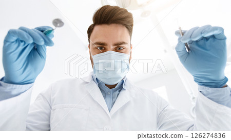 Male dentist in protective face mask and medical gloves holding dental tools, making checkup for patient. Young man stomatologist doing treatment, closeup portrait, low angle shot. 126274836