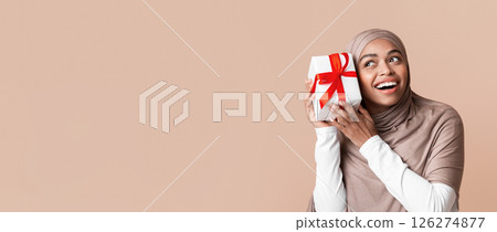 Surprise gift. Joyful afro muslim woman in hijab holding present, shaking whapped box, guessing what's inside, standing over yellow studio background Surprise gift. Joyful afro muslim woman in hijab holding present, shaking whapped box, guessing what's inside, standing over yellow studio background 126274877