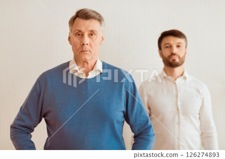 Authoritarian Parent. Serious Mature Son Standing Overshadowed By His Elderly Father Looking At Camera Over White Background. Selective Focus Authoritarian Parent. Serious Mature Son Standing Overshadowed By His Elderly Father Looking At Camera Over White Background. Selective Focus 126274893