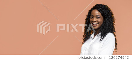 A happy black woman with curly, black hair is smiling while wearing a white shirt. She stands against a peach-colored background and looks directly at the camera. 126274942