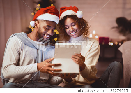 Christmas magic. Afro couple opening gift box in cozy living room, spending evening together 126274965