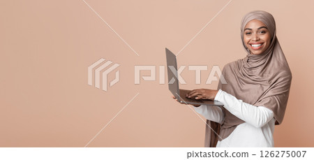 Online education concept. Happy black muslim woman in hijab holding laptop, typing on keyboard and looking at camera, yellow background, free space Online education concept. Happy black muslim woman in hijab holding laptop, typing on keyboard and looking at camera, yellow background, free space 126275007