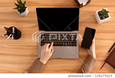Top View Of Office Workplace. Female manager sitting at wooden desk, using laptop and smartphone, mock up, flatlay, copyspace Top View Of Office Workplace. Female manager sitting at wooden desk, using laptop and smartphone, mock up, flatlay, copyspace 126275023
