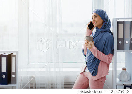 Young muslim businesswoman relaxing in office with smartphone and coffee, standing near window and holding mug with hot drink, copy space Young muslim businesswoman relaxing in office with smartphone and coffee, standing near window and holding mug with hot drink, copy space 126275030