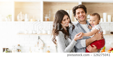This image shows a happy family of three in a modern kitchen. The father is holding their baby, who is smiling and reaching out to the mother 126275041