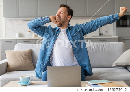A man sits on a couch in a living room, wearing a blue denim shirt and a white t-shirt. He is yawning widely with his arms outstretched. A laptop is open on a table in front of him. 126275042
