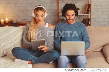 Gadgets. Couple In Headphones Using Laptop And Digital Tablet Sitting On Sofa Indoor. Selective Focus Gadgets. Couple In Headphones Using Laptop And Digital Tablet Sitting On Sofa Indoor. Selective Focus 126275134