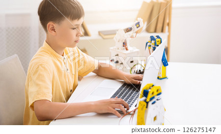 A young boy is learning about robotics by using a laptop computer. The boy is sitting at a table and is looking at the screen of the laptop. There is a robotic arm behind the laptop. 126275147
