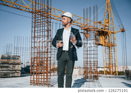 In hard hat, holding plan in hands. Businessman in formal clothes is on the construction site at daytime 126275191