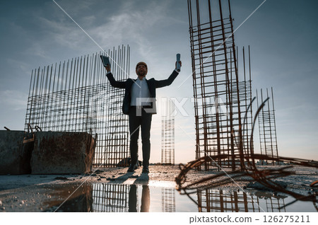 With hands up. Standing at evening time. Businessman in formal clothes is on the construction site With hands up. Standing at evening time. Businessman in formal clothes is on the construction site 126275211