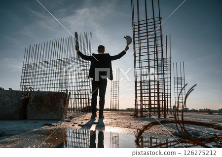 With hands up. Standing at evening time. Businessman in formal clothes is on the construction site 126275212