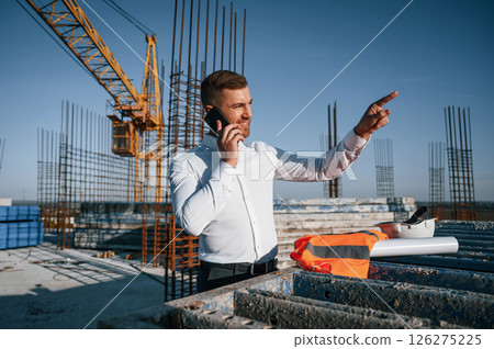 Having conversation by phone. Businessman in formal clothes is on the construction site at daytime Having conversation by phone. Businessman in formal clothes is on the construction site at daytime 126275225