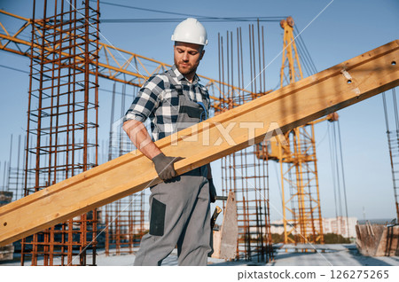 With big wooden plank in hand. Man is working on the construction site at daytime With big wooden plank in hand. Man is working on the construction site at daytime 126275265