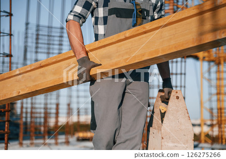 With big wooden plank in hand. Man is working on the construction site at daytime With big wooden plank in hand. Man is working on the construction site at daytime 126275266