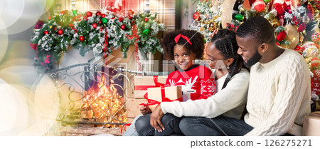 Christmas gift from parents. Cute little african girl holding Xmas present, sitting with her mom and dad in decorated living room 126275271