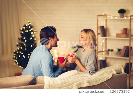 Young couple celebrating Christmas at home, relaxing with cups of coffee on sofa 126275302
