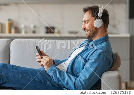 A man wearing headphones is sitting on a grey couch, smiling while looking at his smartphone. He has a white t-shirt underneath the denim shirt and his legs are stretched out in front of him. A man wearing headphones is sitting on a grey couch, smiling while looking at his smartphone. He has a white t-shirt underneath the denim shirt and his legs are stretched out in front of him. 126275328