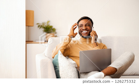 Positive Afro Man Using Laptop Talking On Mobile Phone And Laughing Sitting On Sofa Indoor. Selective Focus, Copy Space 126275332