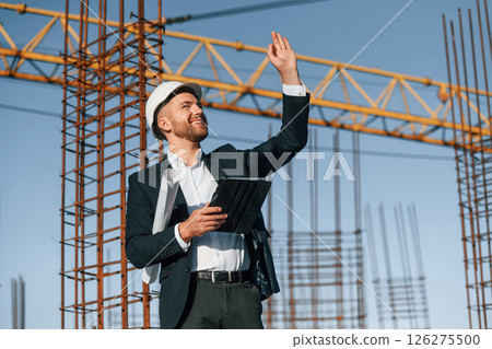 Giving orders by hand gesture. Businessman in formal clothes is on the construction site at daytime Giving orders by hand gesture. Businessman in formal clothes is on the construction site at daytime 126275500