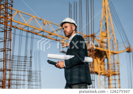 Side view. Walking and holding paper with plan and digital tablet. Businessman in formal clothes is on the construction site Side view. Walking and holding paper with plan and digital tablet. Businessman in formal clothes is on the construction site 126275507