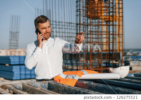 Standing and talking by phone. Businessman in formal clothes is on the construction site at daytime Standing and talking by phone. Businessman in formal clothes is on the construction site at daytime 126275521