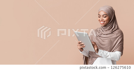 Portrait of smiling afro muslim girl in hijab using digital tablet, downloading new app or browsing media, standing over yellow background, free space 126275610
