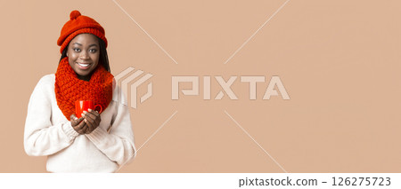 African American woman wearing a knitted red hat and a large scarf smiles while holding a cup. The warm colors of her outfit and the soft background create a cozy atmosphere, perfect for chilly days. 126275723