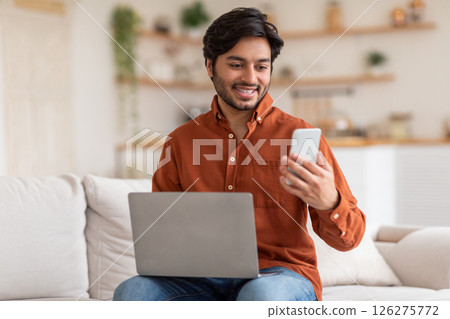 Arab man sits on a white couch in a living room. He is working on a laptop while looking at his phone. He is smiling, suggesting he is enjoying his work. 126275772