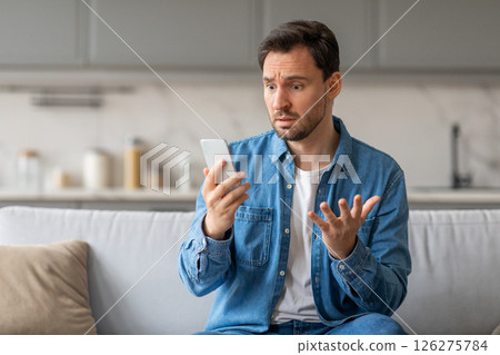 A man sits on a light gray couch in a kitchen, staring at his smartphone with a confused expression. He has his right hand raised, gesturing with his fingers, while holding the phone in his left hand. 126275784