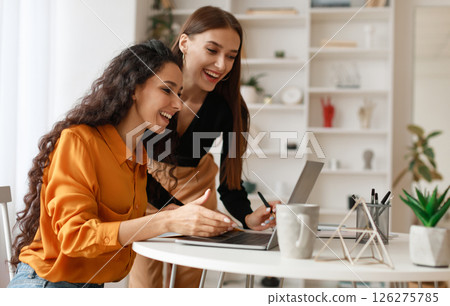 Two Happy Ladies Using Laptop Computer Working And Learning Online Together Or Making Video Call Sitting At Desk In Modern Office. E-Learning. Females Entrepreneurship Career Concept. Side View 126275785