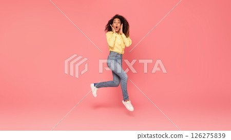 African American young woman with dark curly hair jumps in the air with her hands on her cheeks. She is looking at the camera with a surprised expression. The background is a bright pink. 126275839
