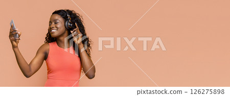 African American young woman with curly dark hair, wearing a coral sleeveless top, smiling and listening to music with headphones while holding a smartphone in her right hand 126275898
