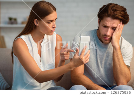 Infidelity. Jealous Girlfriend Showing Her Cheating Boyfriend His Phone Demanding Explanation Sitting On Sofa Indoor 126275926