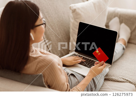A woman sits on a couch, using her laptop and credit card to shop online. 126275973