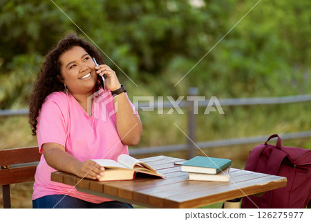 A young woman sits at a wooden table in a park, enjoying a phone conversation. She has a book open in front of her, with several other books nearby, surrounded by greenery on a sunny day. A young woman sits at a wooden table in a park, enjoying a phone conversation. She has a book open in front of her, with several other books nearby, surrounded by greenery on a sunny day. 126275977