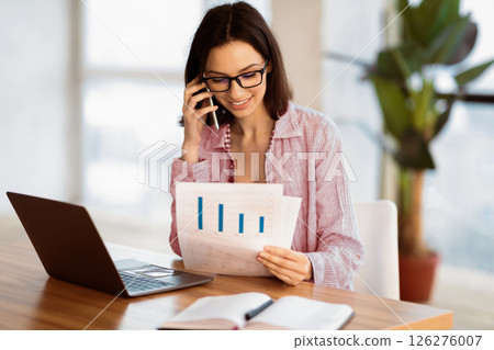 Business, Technology And Communication Concept. Young woman making phone call while looking at document with graphs 126276007