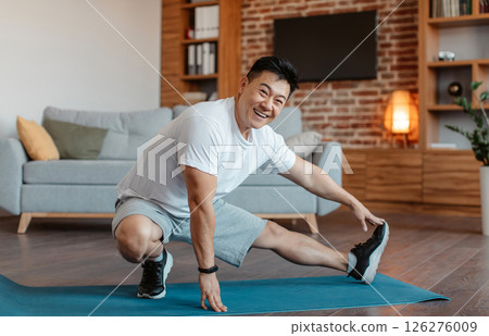 Happy mature sportsman making exercises for legs, stretching on fitness mat in living room interior and smiling at camera. Home training, workout, fitness and weight loss 126276009