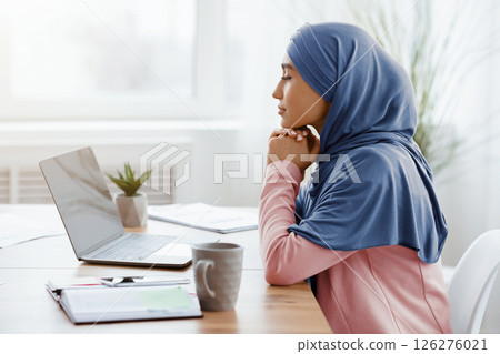 Business Planning. Profile portrait of thoughtful arabic female entrepreneur sitting at workplace in office and looking at laptop screen, side view Business Planning. Profile portrait of thoughtful arabic female entrepreneur sitting at workplace in office and looking at laptop screen, side view 126276021