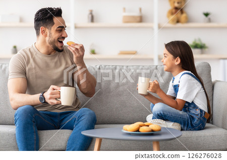 Snack Time. Portrait of cheerful little girl and man holding cups, drinking coffee and tea, eating cookies and looking at each other, dad and daughter talking sitting on the couch in living room Snack Time. Portrait of cheerful little girl and man holding cups, drinking coffee and tea, eating cookies and looking at each other, dad and daughter talking sitting on the couch in living room 126276028